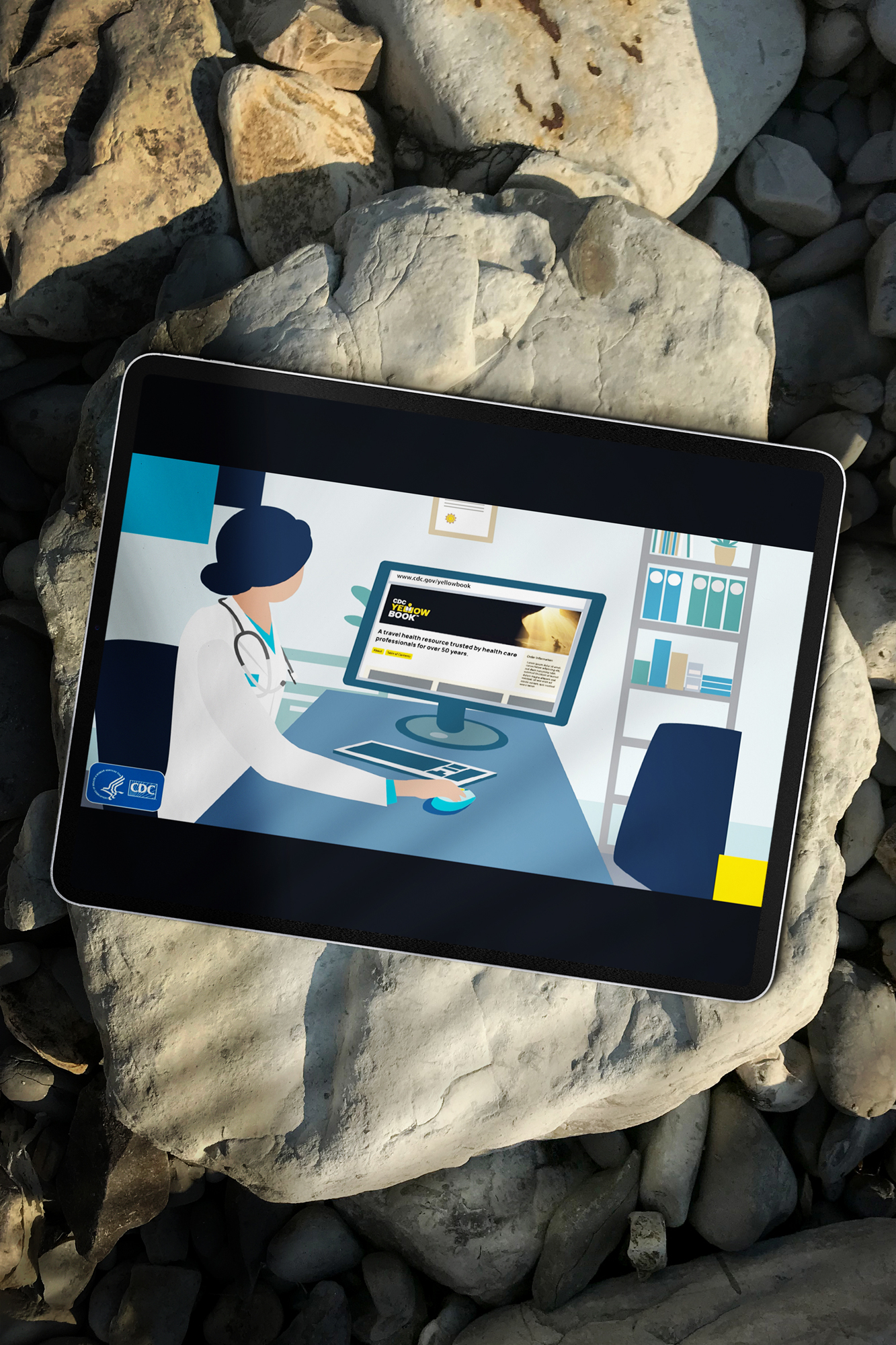 A tablet propped against rocks showing a Yellow Book web banner: an illustrated clinician at a desk with the book open on their monitor.