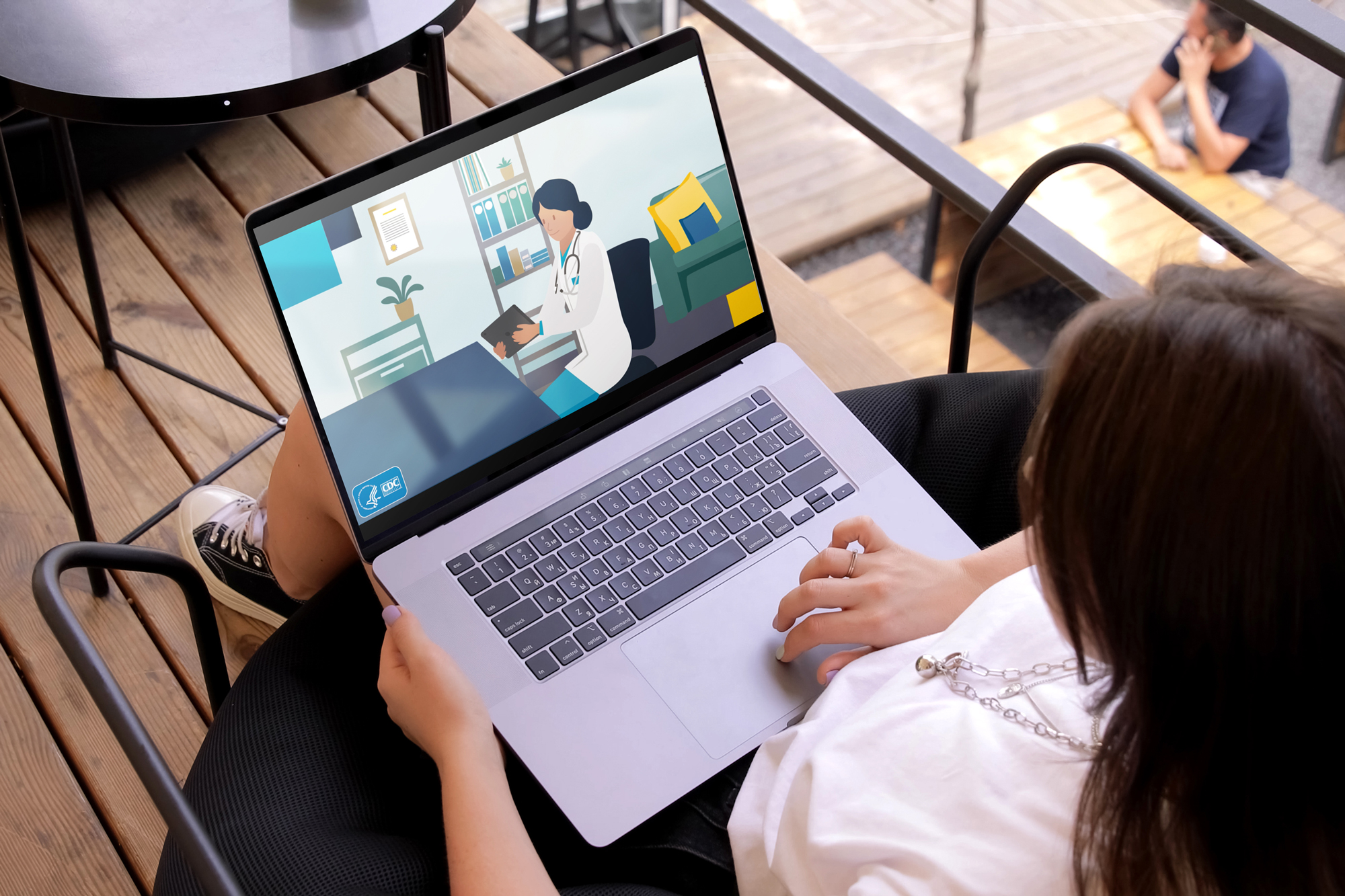 A woman at an outdoor cafe table reading an illustrated CDC Yellow Book web page on a laptop.