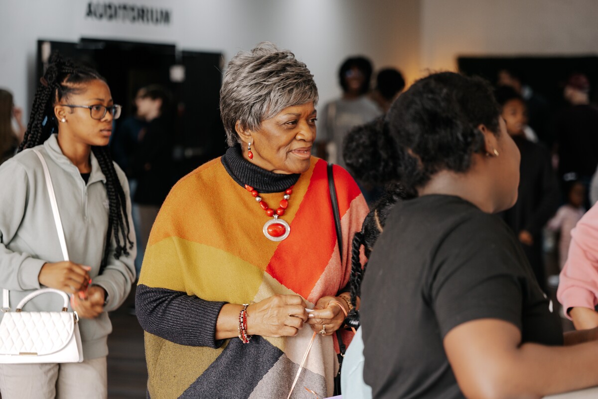Older woman wearing a colorful poncho and red jewelry smiling and interacting with a group of people indoors near an auditorium entrance.