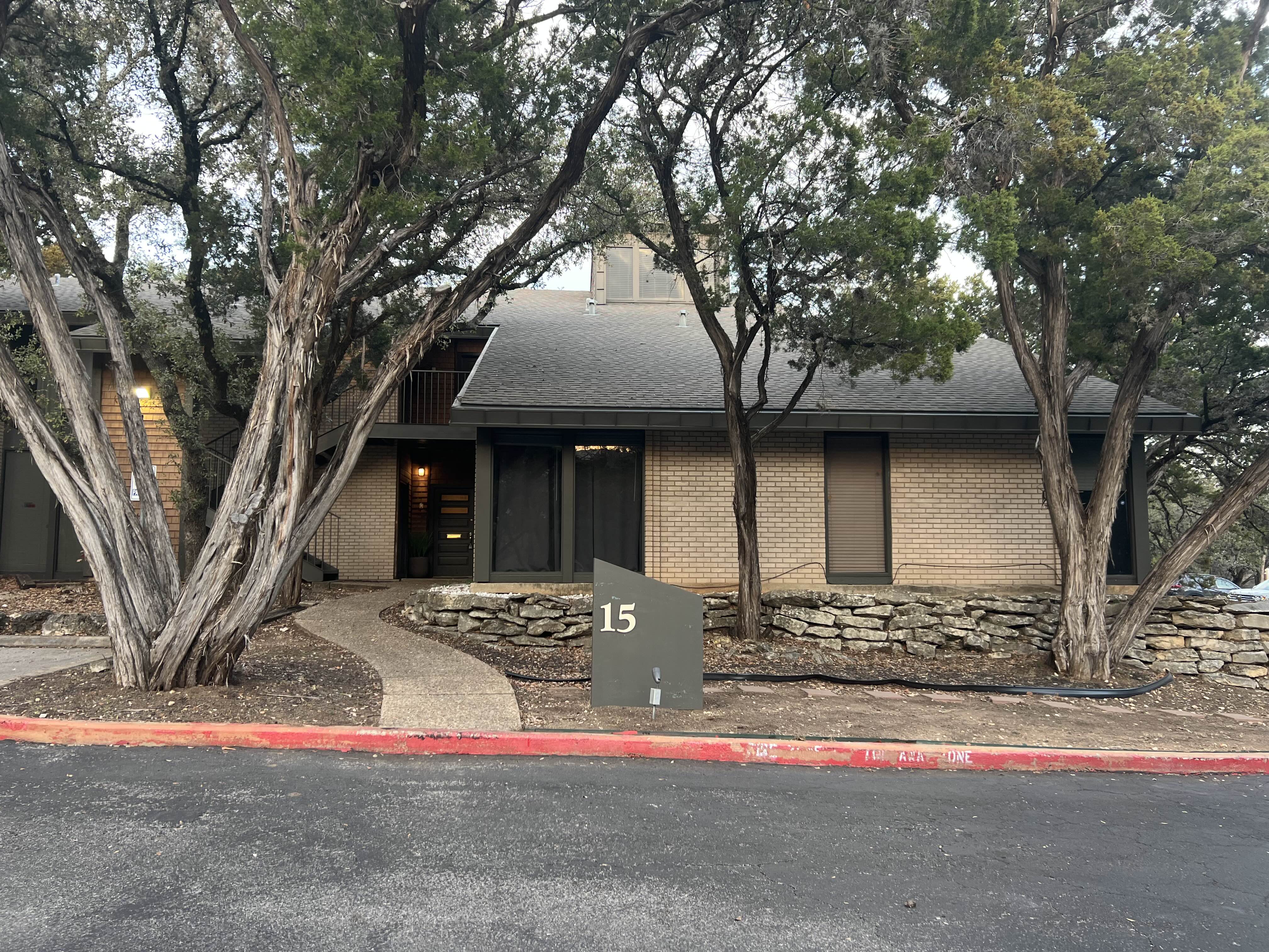 Building with beige brick wall, dark framed windows, entrance door, surrounded by stone landscaping and large trees, with a dark green sign numbered 15 in front.