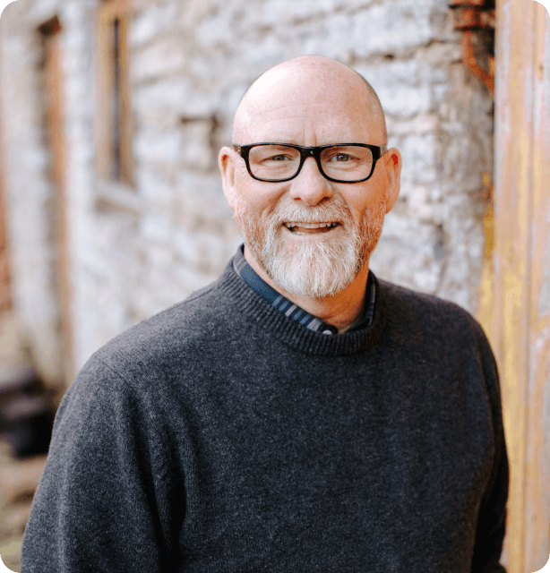 Man with glasses and a beard smiling at the camera. In the background is a red brick house.