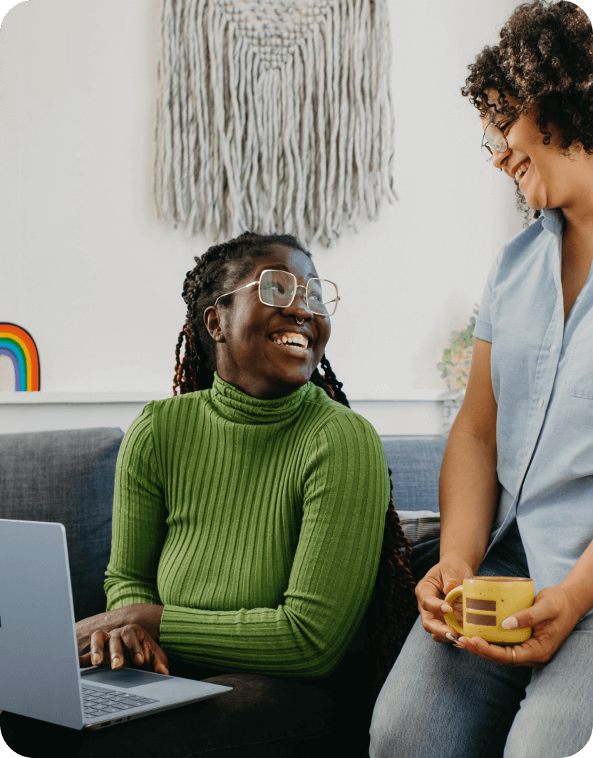 2 people smiling at each other on a sofa. One black person is on a laptop.