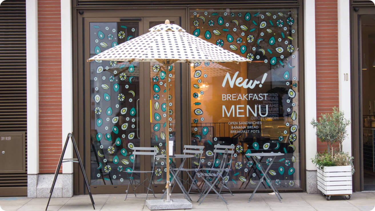 Detox KItchen store windows full of illustrations of eggs, avocados, bananas, lettuce and the text on the window reads "New breakfast menu - open sandwiches, banana bread, breakfast pots"