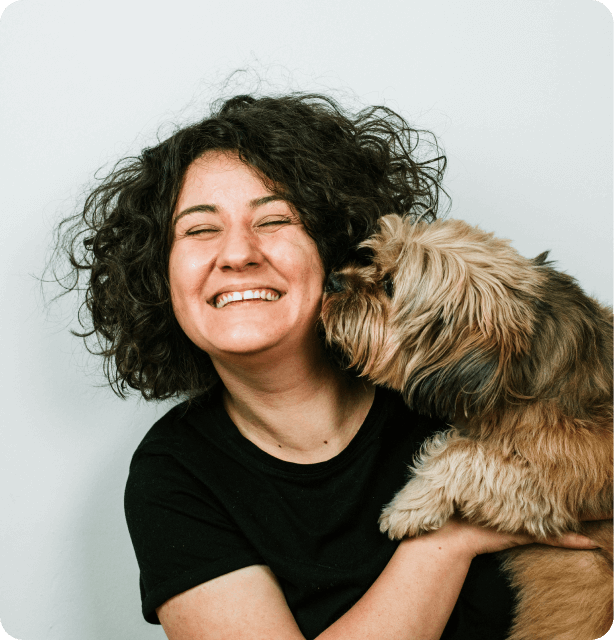 Lady with very dark curly hair smiling while a dog she is cuddling sniffs her cheek.