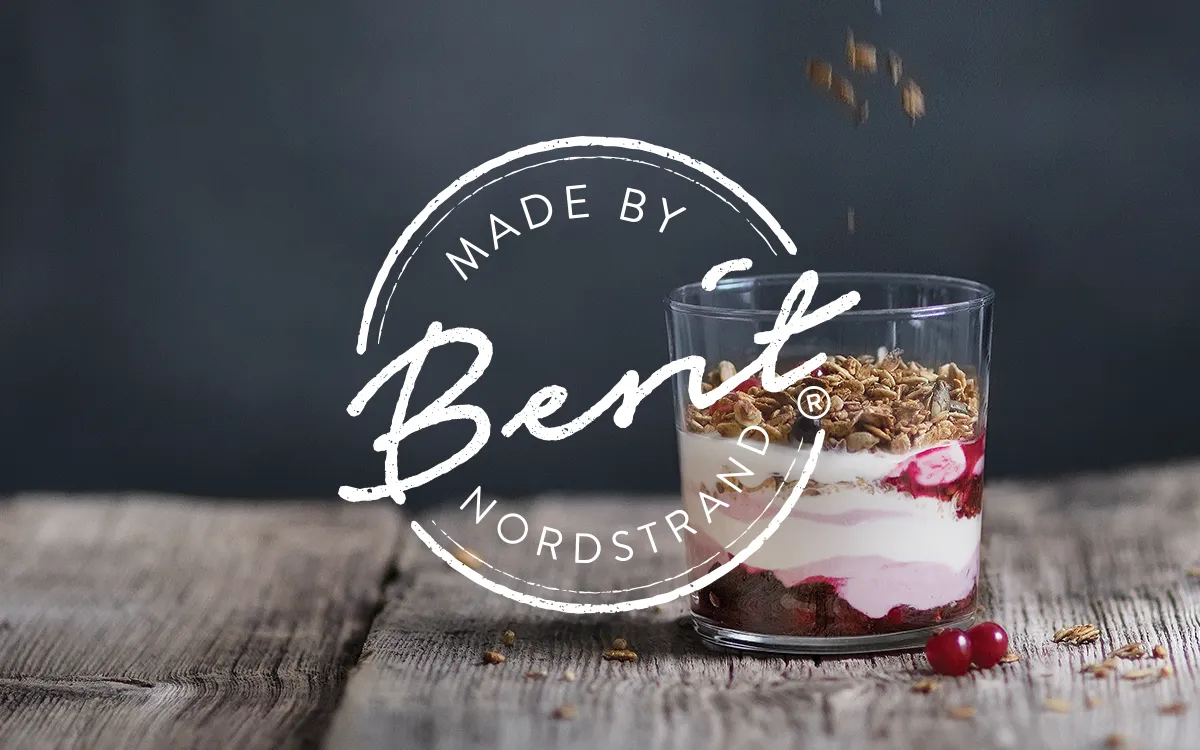 Et glass med lagdelt yoghurt og müsli på en treflate, med noen røde bær og dryss av müsli i luften. Berit Nordstrand logoen