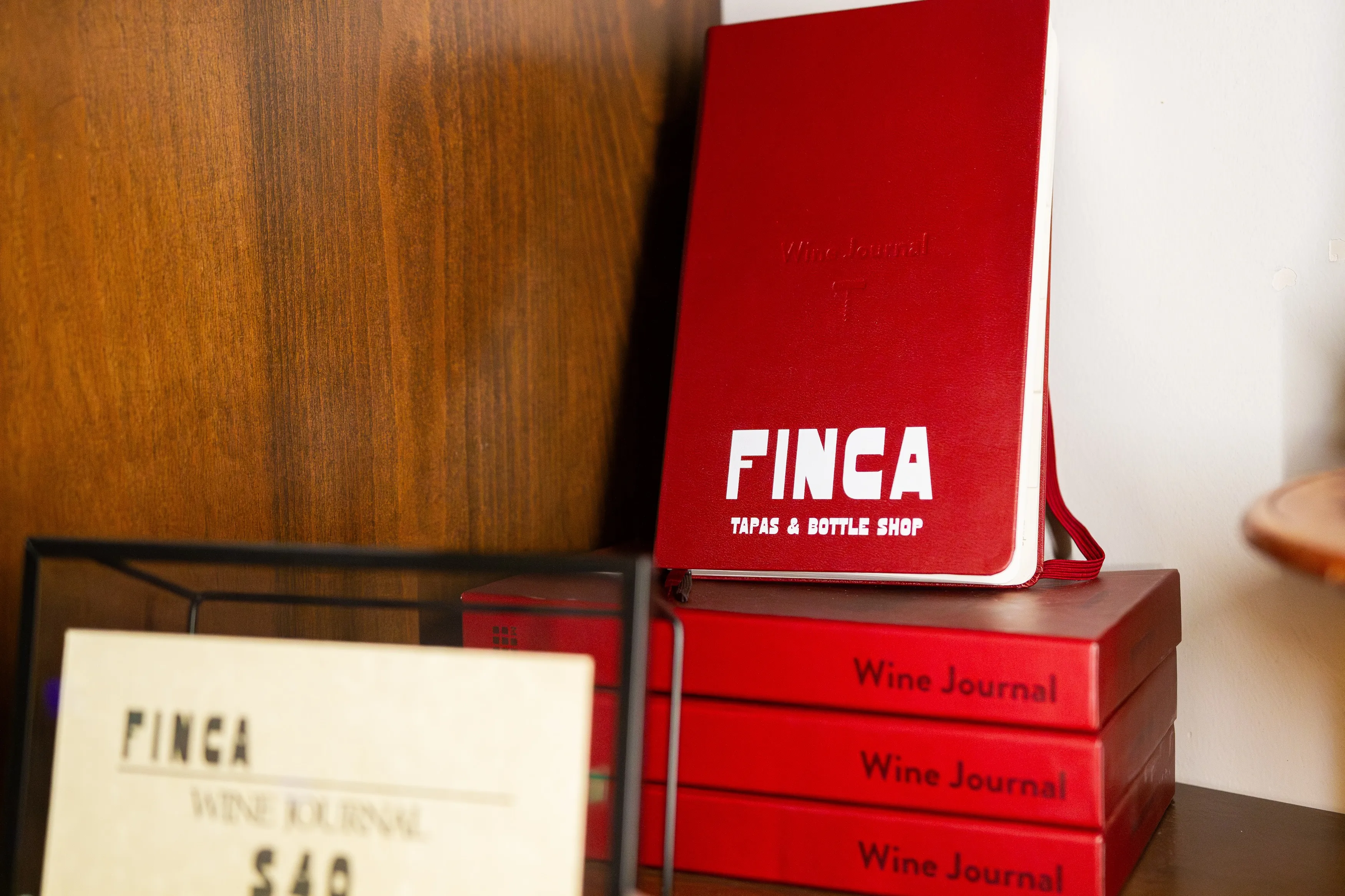 Stack of three red Wine Journal books and a red notebook labeled 'FINCA TAPAS & BOTTLE SHOP' on a wooden shelf.