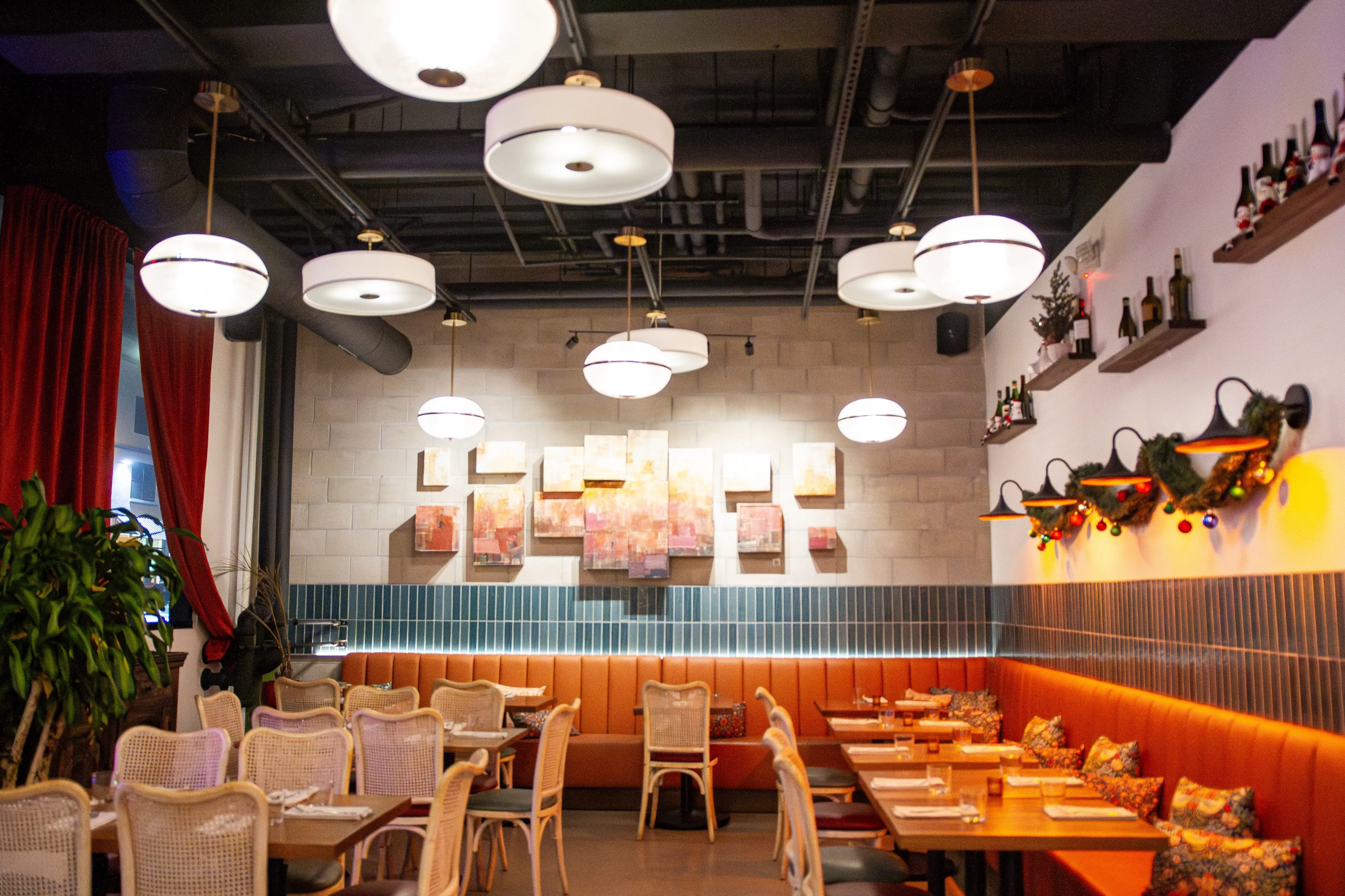 Cozy restaurant interior with orange banquette seating, wooden tables, rattan chairs, decorative pillows, hanging globe lights, and wall art.