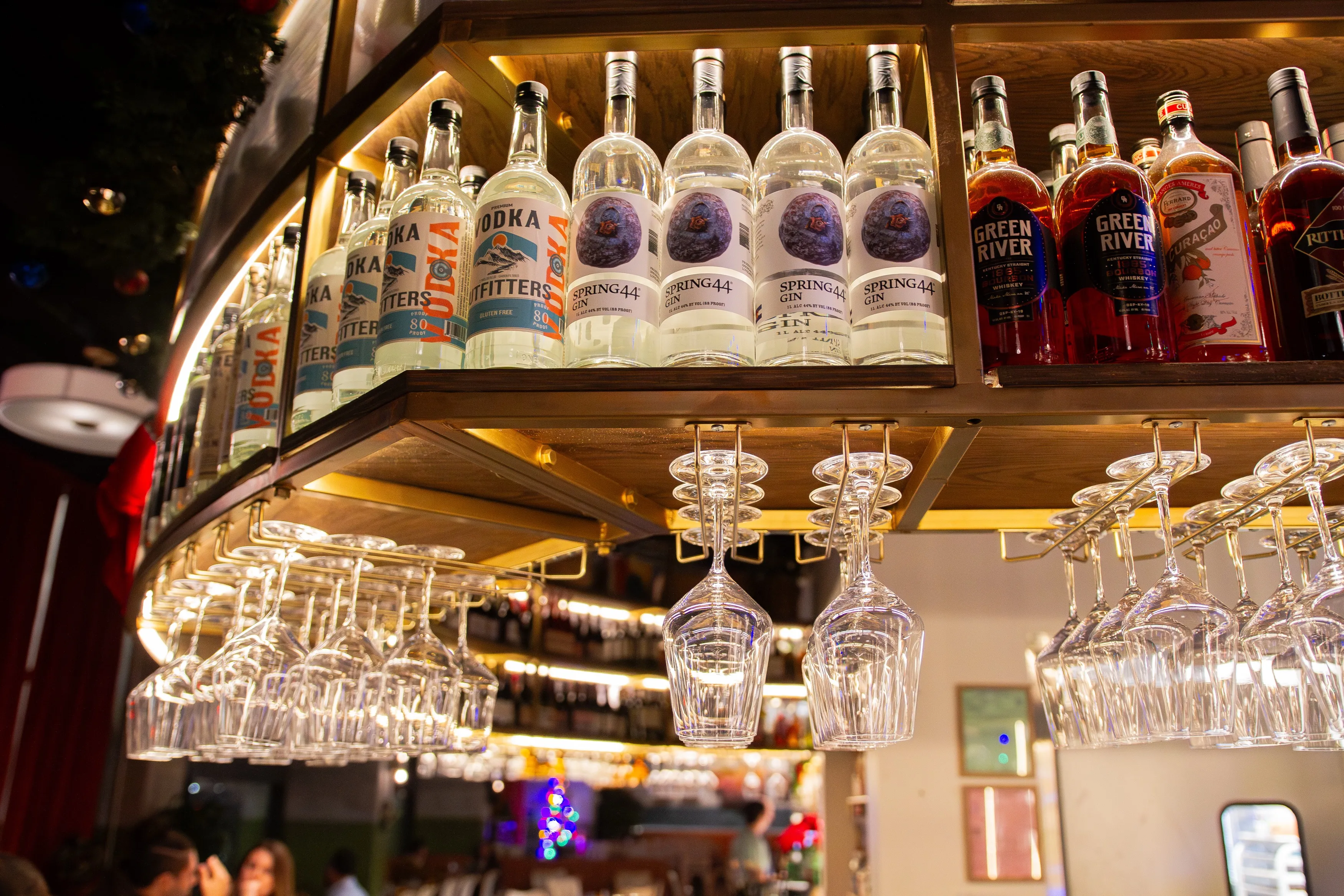 Bottles of vodka, gin, and whiskey on a wooden bar shelf with wine glasses hanging upside down below.