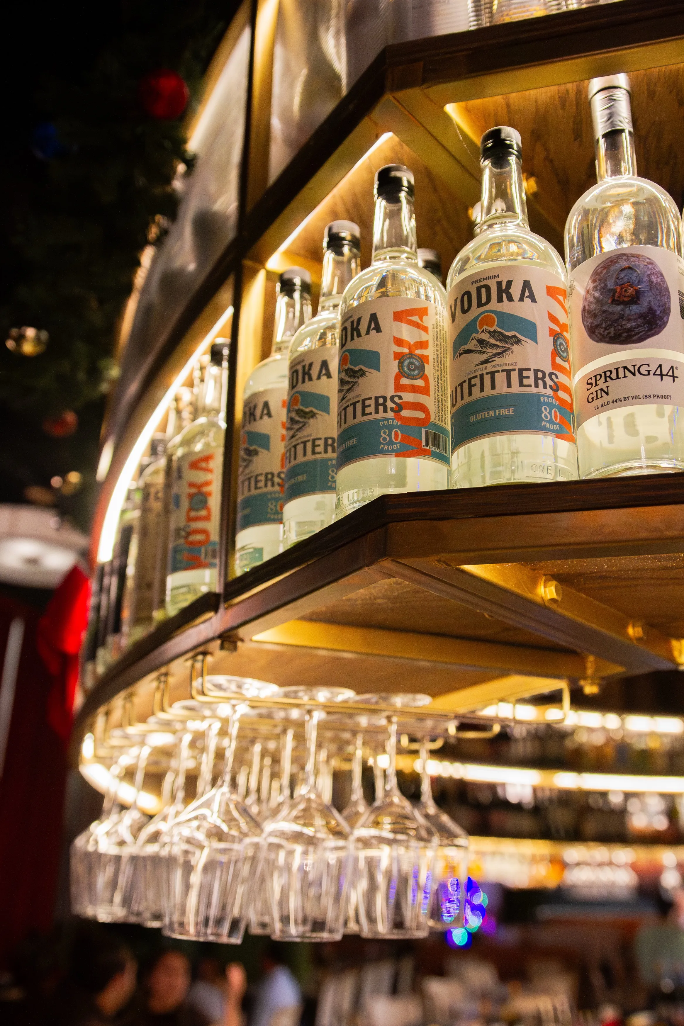 Bottles of Utah Vodka and Spring44 Gin displayed on a backlit wooden shelf above hanging wine glasses in a bar.