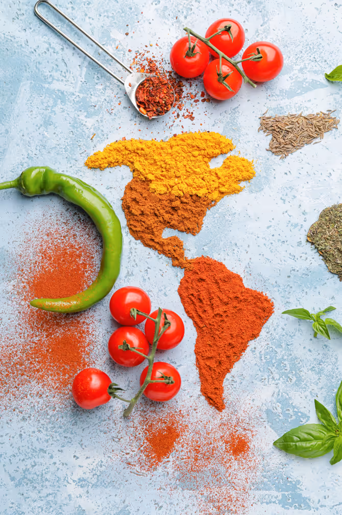 Map of the Americas made with yellow, brown, and red spices surrounded by fresh tomatoes, a green chili pepper, and various herbs and spices on a blue surface.