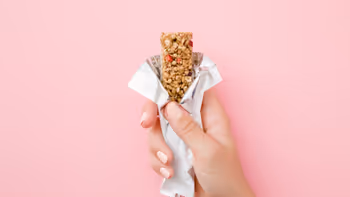 Hand holding a partially unwrapped granola bar against a pink background.
