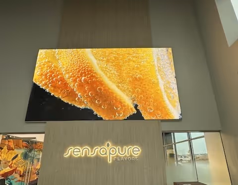 Large digital screen displaying a close-up image of orange slices with bubbles above the illuminated Sensapure Flavors logo inside a modern building.