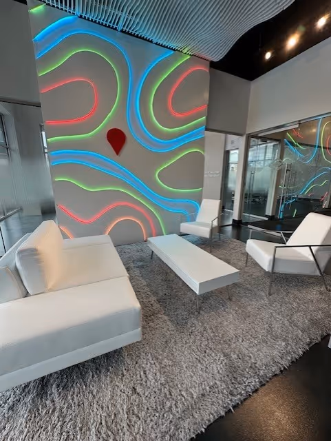 Modern lounge area with white sofas and chairs, a low white coffee table, gray shag carpet, and a wall decorated with colorful neon lines and a red teardrop shape.