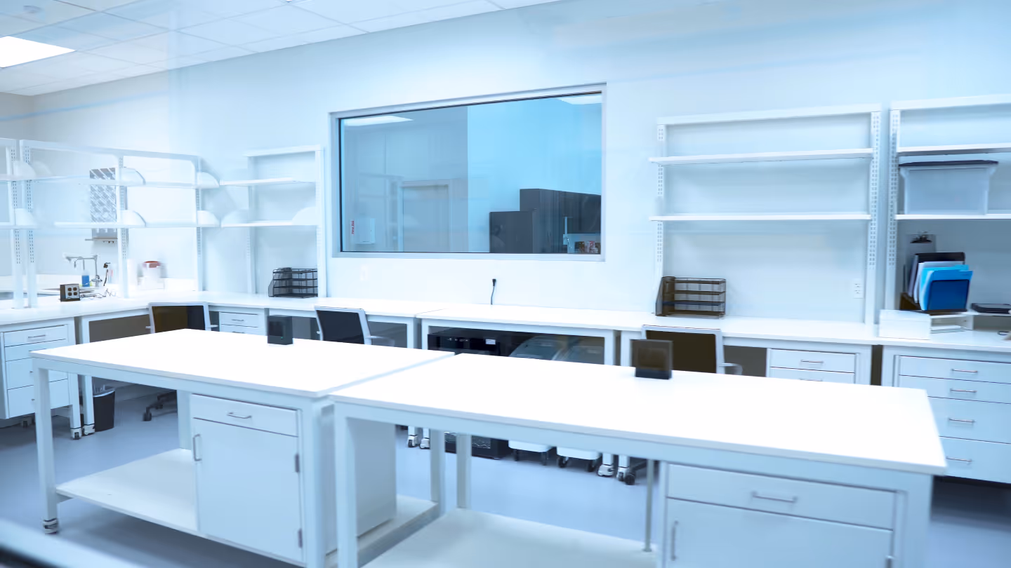Bright, clean laboratory workspace with white tables, chairs, shelves, and cabinets under soft lighting.