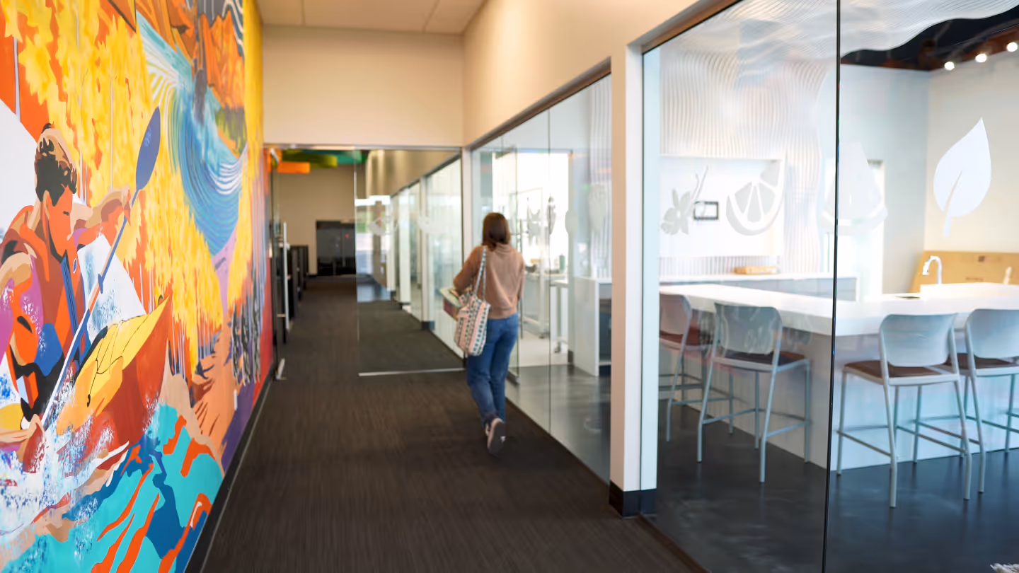 Person walking down a carpeted hallway with a colorful mural on the left and glass-walled office on the right.