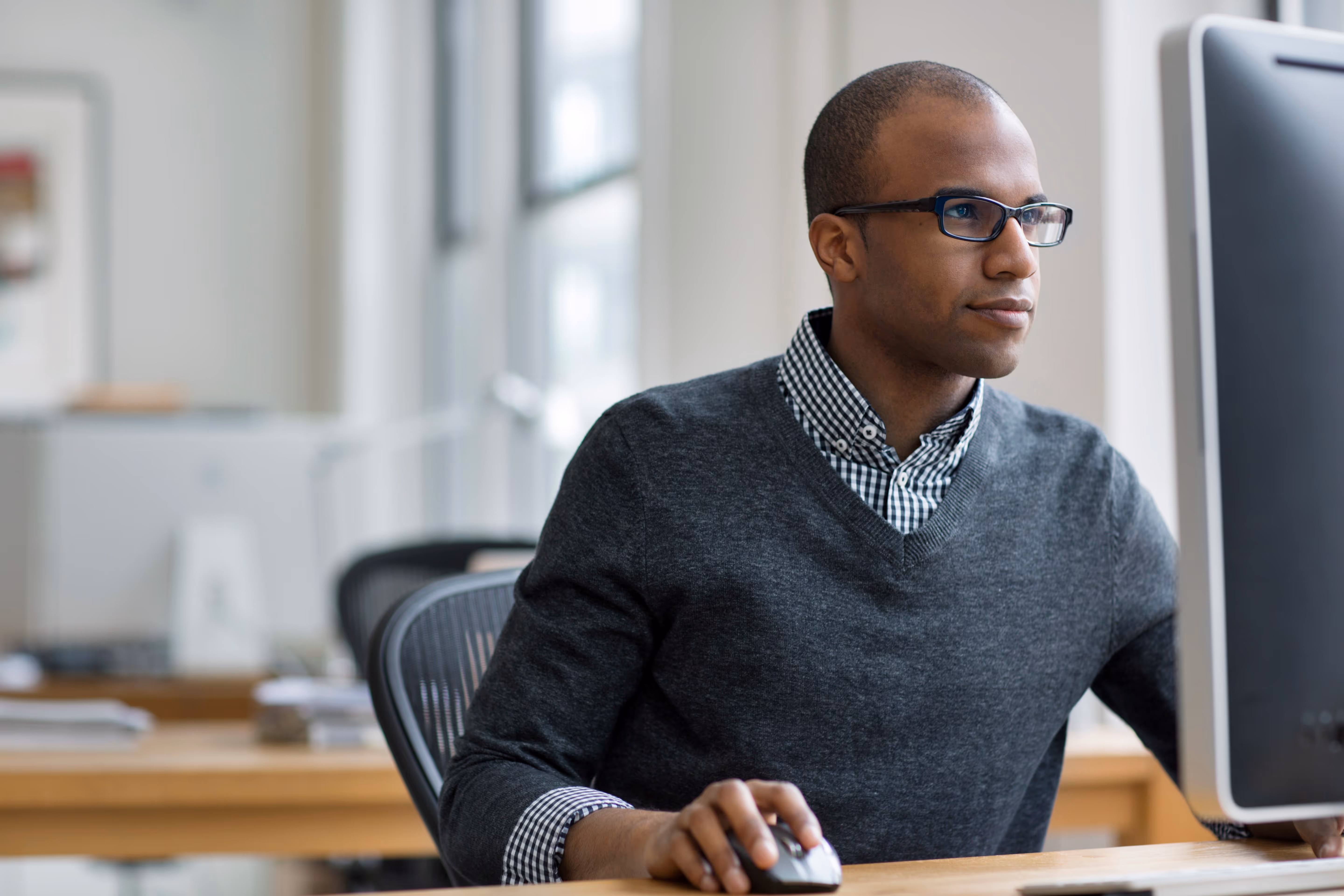 Man wearing glasses and a gray sweater using a desktop computer in an office setting.