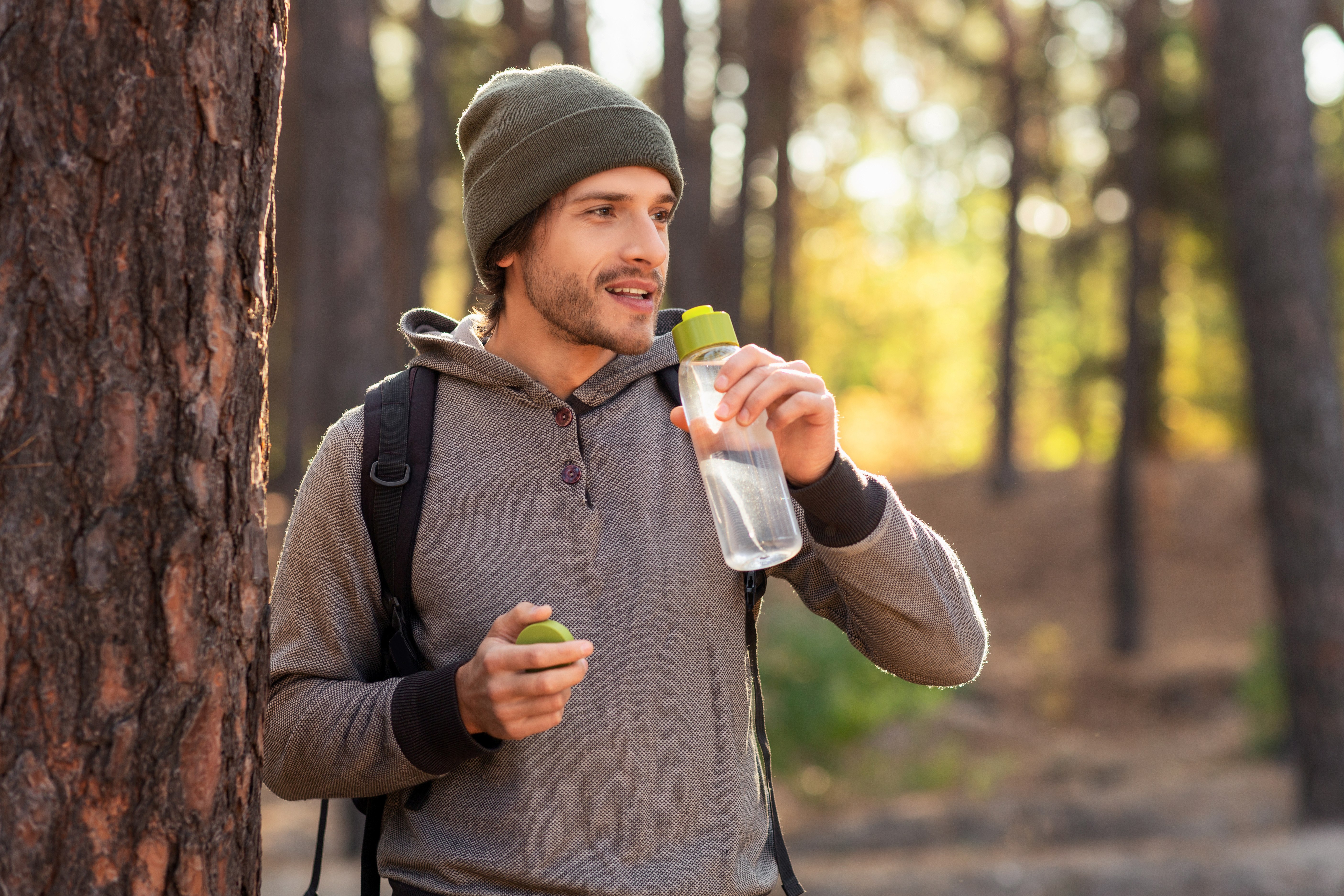 Young man wearing a beanie and hoodie leaning against a tree in a forest, drinking water from a clear bottle with a green cap.