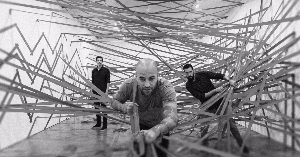 Three men navigating through a dense web of tape strips stretched across a white gallery space. The strips zigzag in patterns resembling financial line charts. One man in a t-shirt pushes through the tape in the foreground while two others move through the