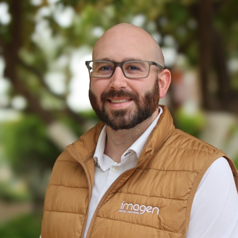 Smiling bald man with glasses and beard wearing a brown vest over a white shirt, standing outdoors with blurred greenery in the background.