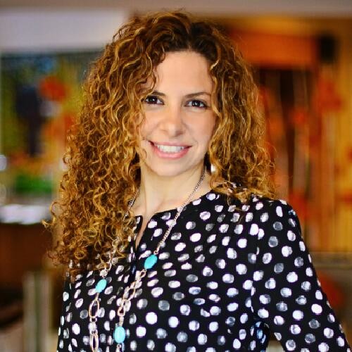 Smiling woman with curly hair wearing a black and white polka dot blouse and a turquoise necklace in an indoor setting.