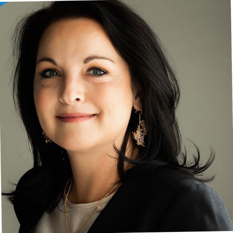Smiling woman with dark hair wearing earrings, a light top, and a dark jacket against a neutral background.