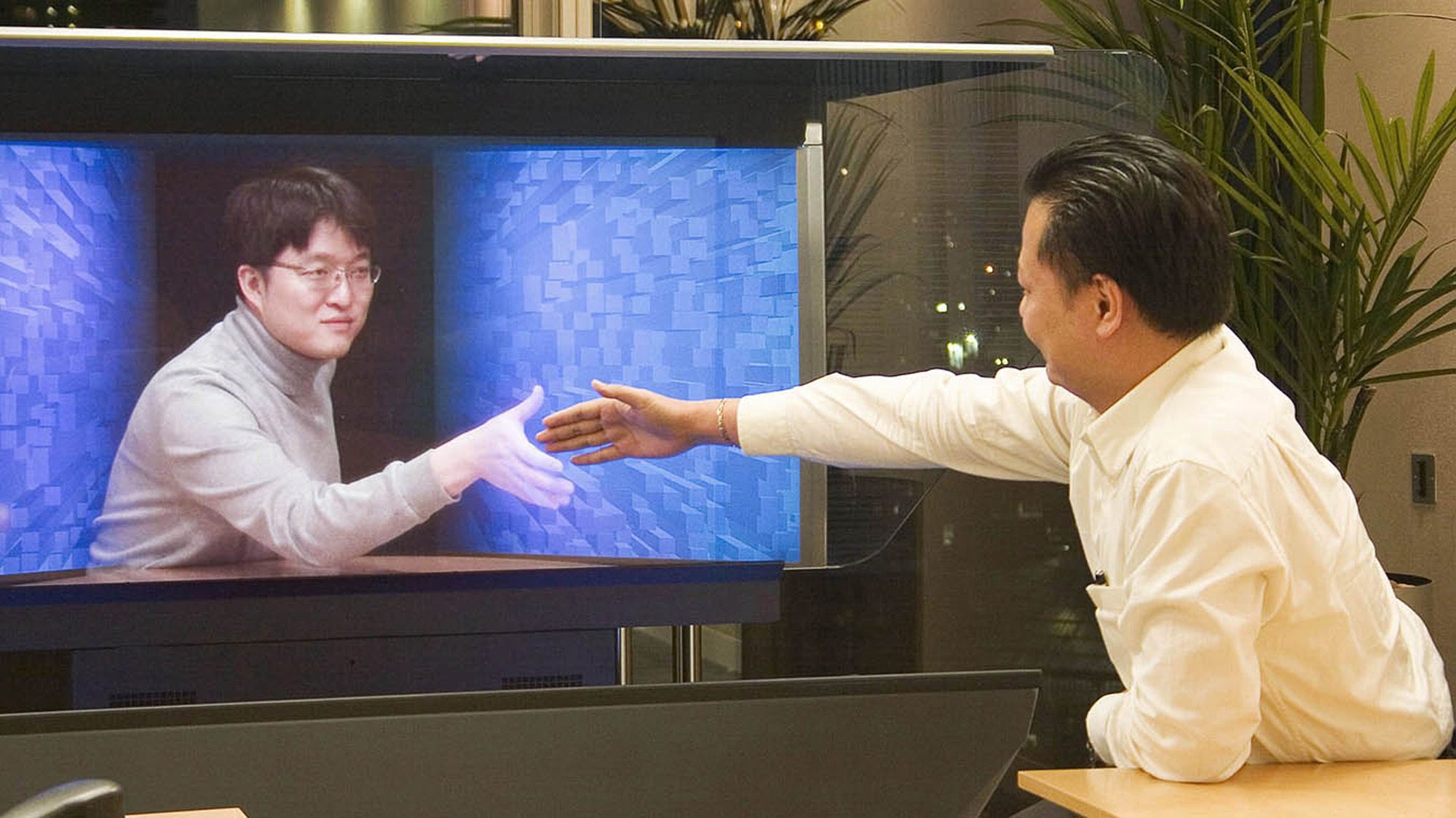In a 1980s-era advertising photo, two businessmen reach out to shake hands. One man is in a potted plant-filled office, and the other, a slightly translucent hologram, reaches out to him from a futuristic-looking monitor.