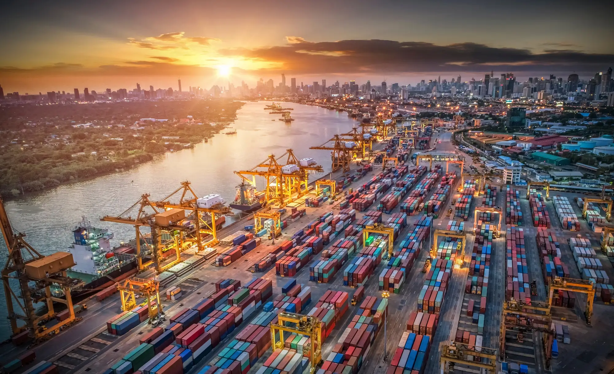 Aerial view of a busy shipping port with rows of colorful containers and cranes alongside a river at sunset.