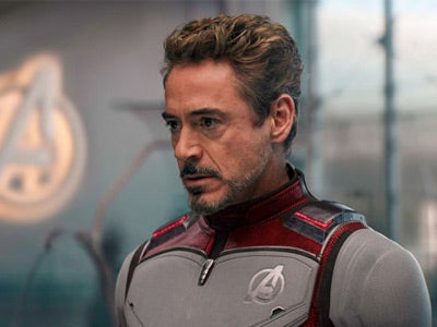 Man in a gray and red superhero suit stands indoors with a focused expression. An "A" logo is visible in the blurred background.