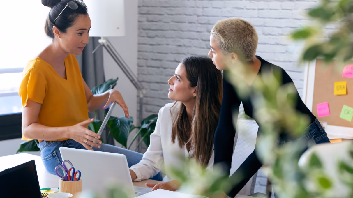 Three women engaged in a discussion around a laptop in a bright modern office space.