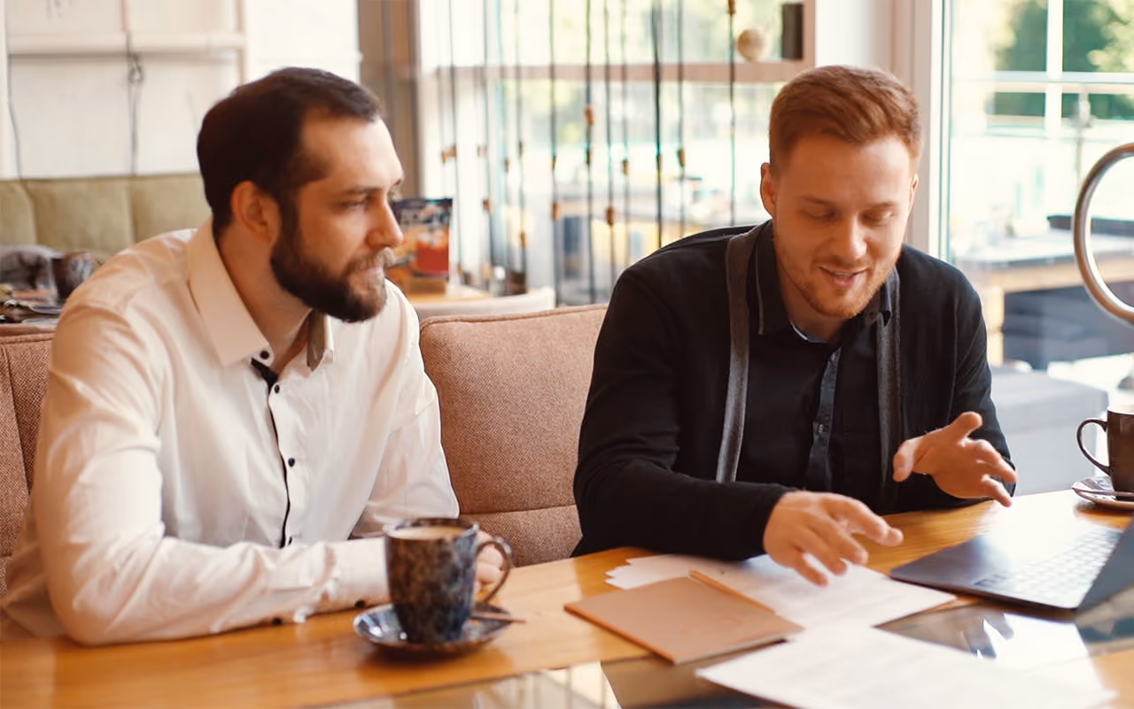 Two men sit at a table with documents and laptops, having a discussion over coffee.