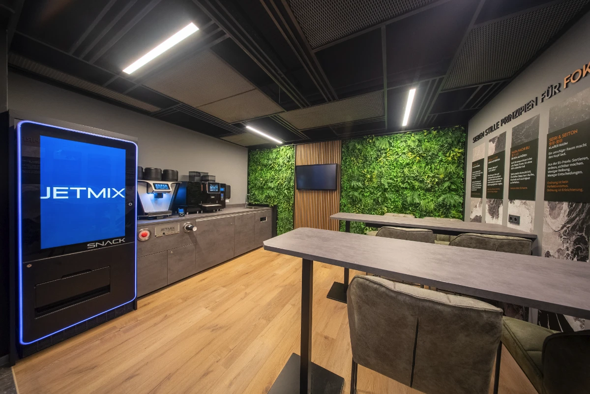 Modern break room with snack vending machine, coffee machines, wooden tables, cushioned chairs, and green plant wall panels.