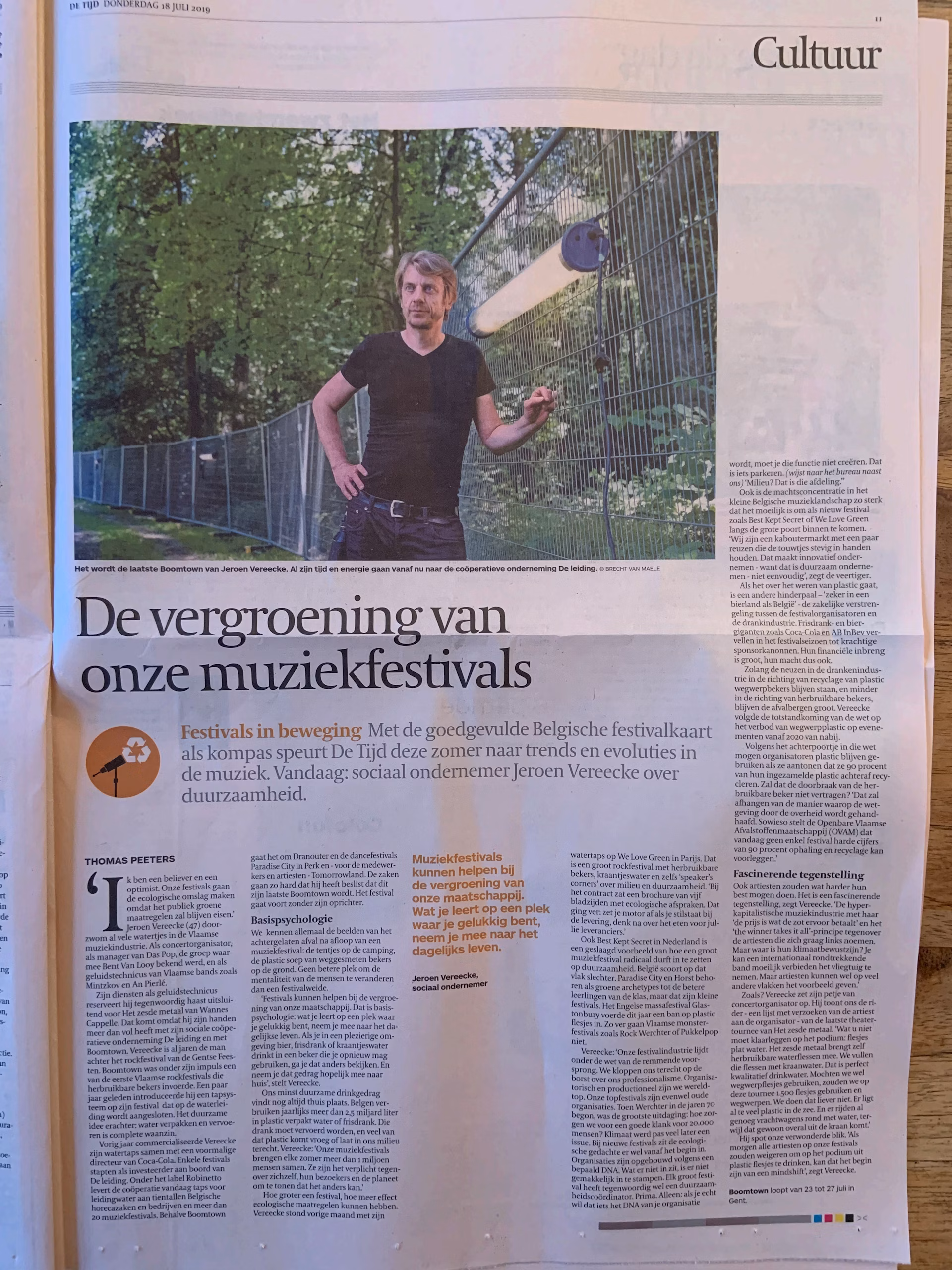 Een opengevouwen krant met een artikel over de verduurzaming van muziekfestivals. Bovenaan een grote foto van Jeroen Vereecke, oprichter van Robinetto, die bij een hek in een groene omgeving staat.