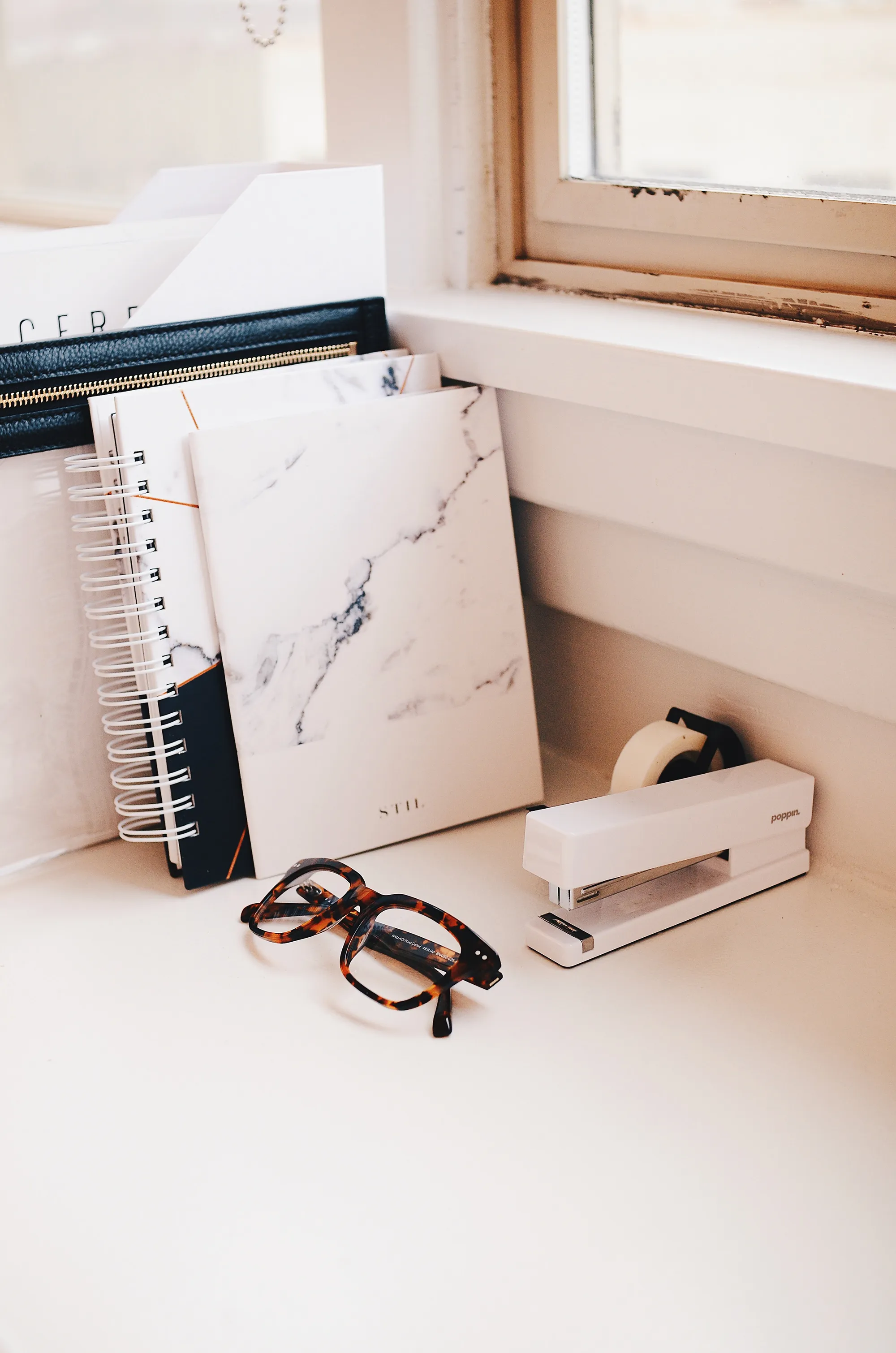 Notebooks, a pencil, and eyeglasses on a desk beside a window.
