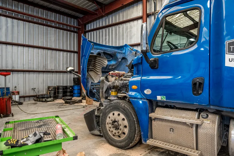 Engine diagnostics: blue day-cab semi with hood up, engine exposed beside creeper cart, oil drums; door decal visible.
