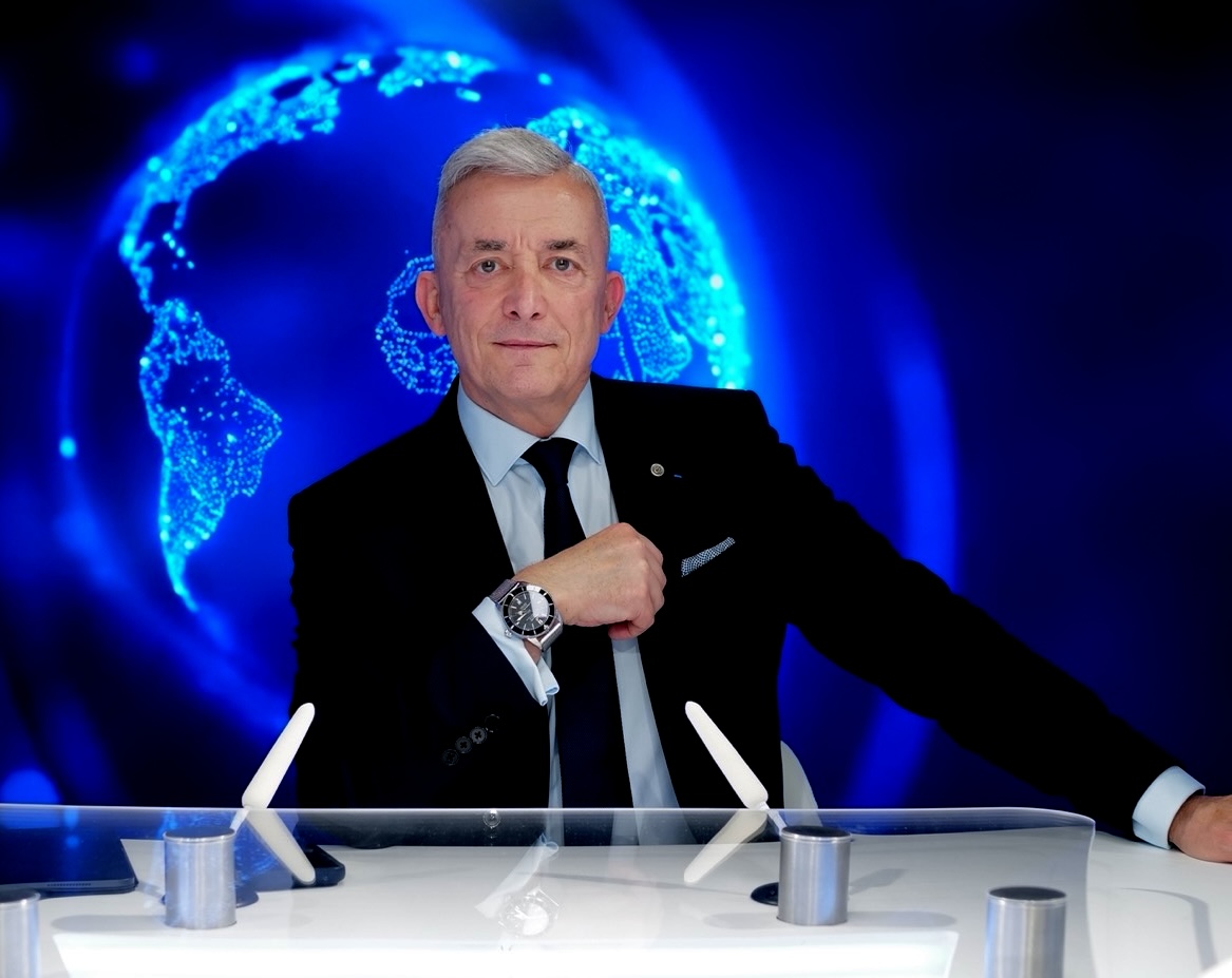 Laurent Vibert  en costume noir avec une cravate noire, assis devant un mur avec une image lumineuse de la Terre en bleu.