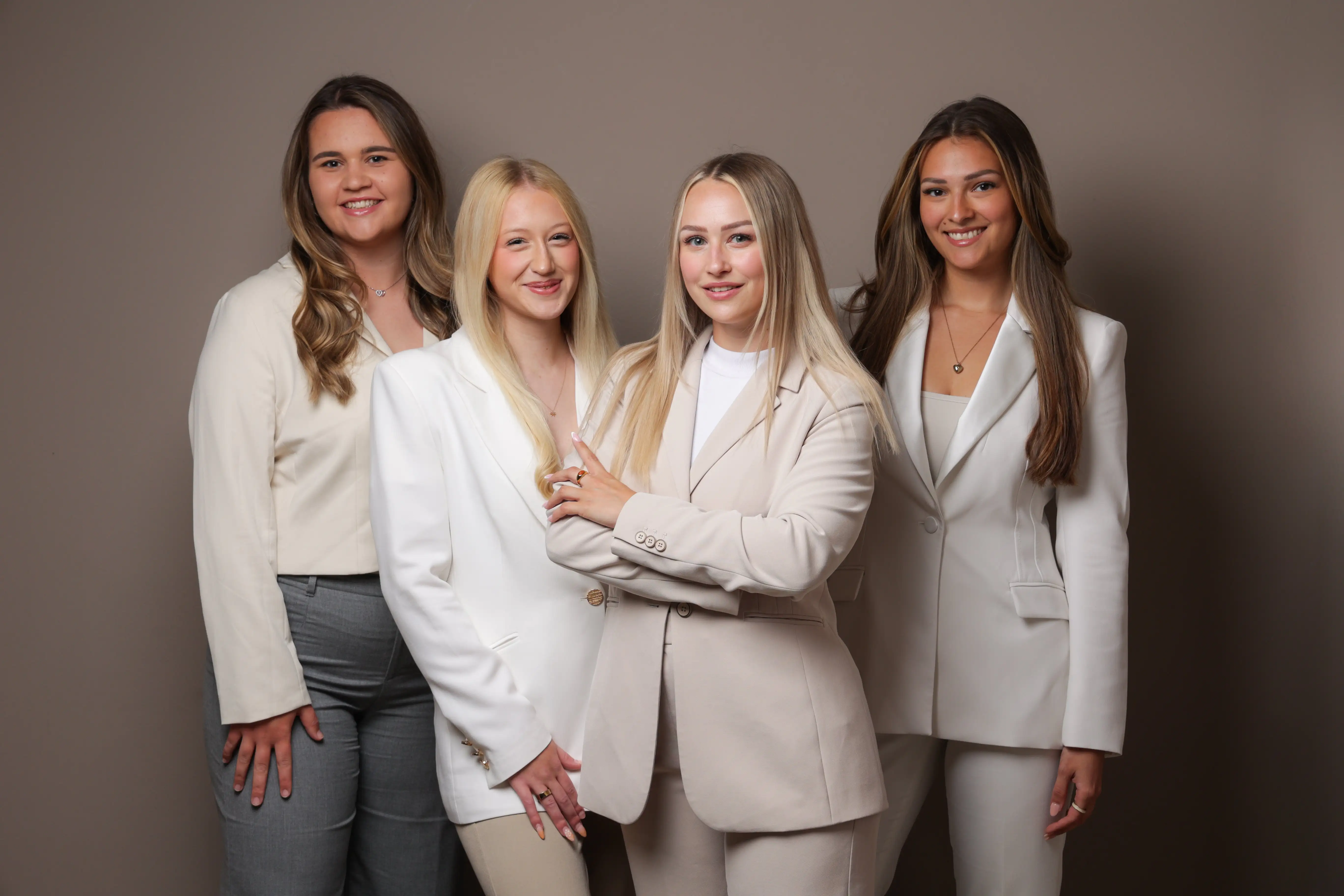 Four women in business attire standing together against a plain neutral background, smiling.