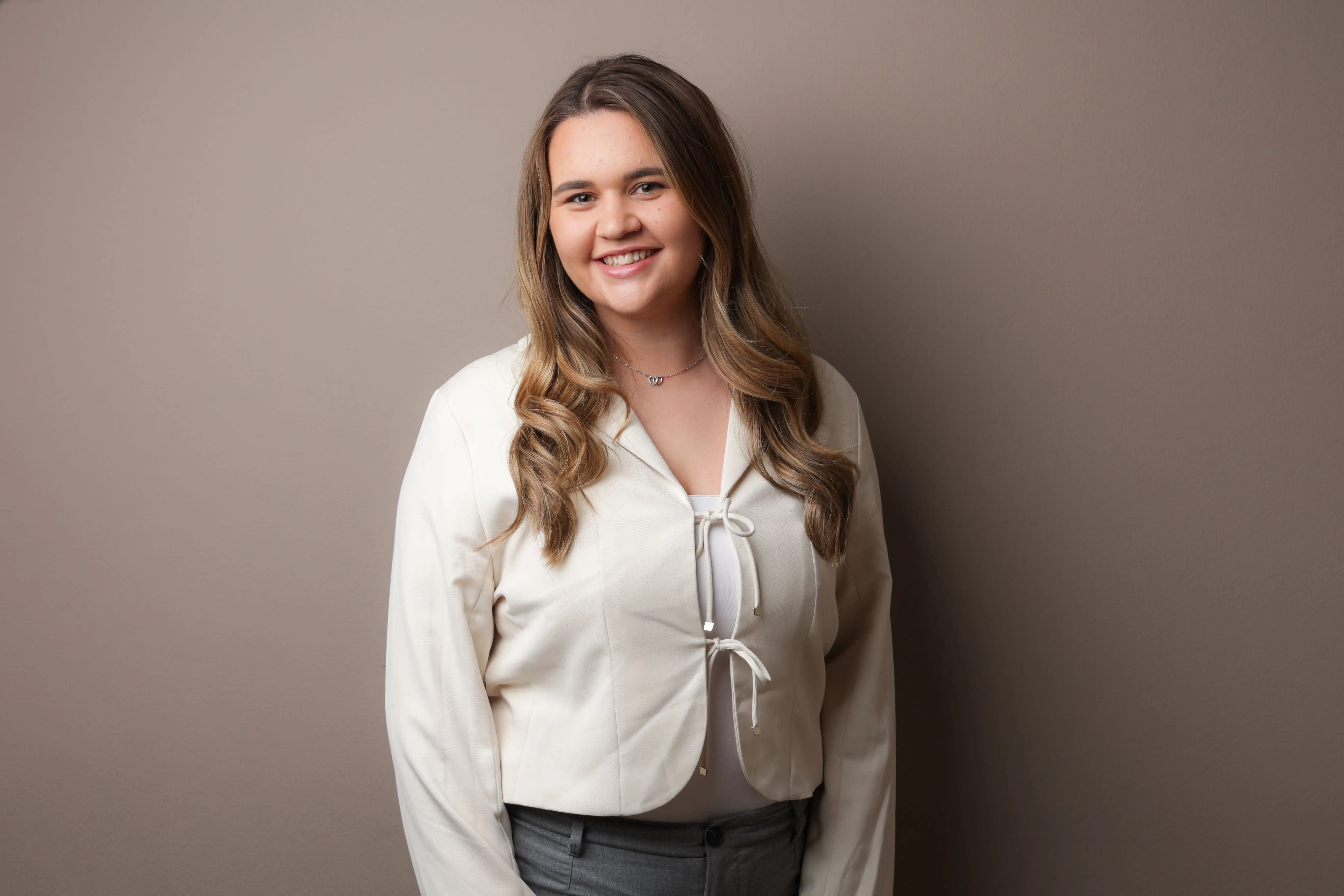 Smiling woman with long wavy hair wearing a white jacket and gray pants against a plain wall.
