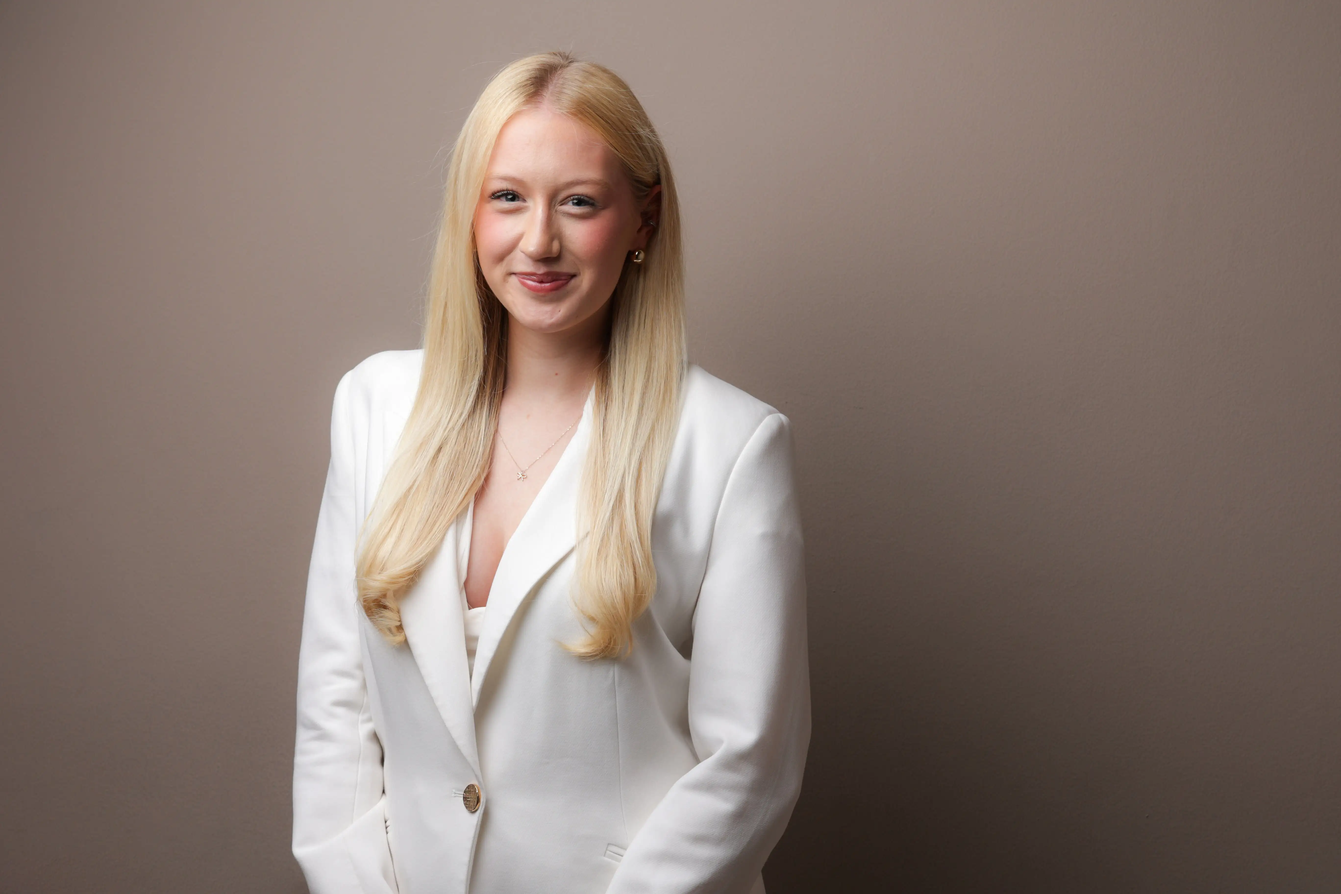 Blonde woman wearing a white blazer smiling with a plain brown background.