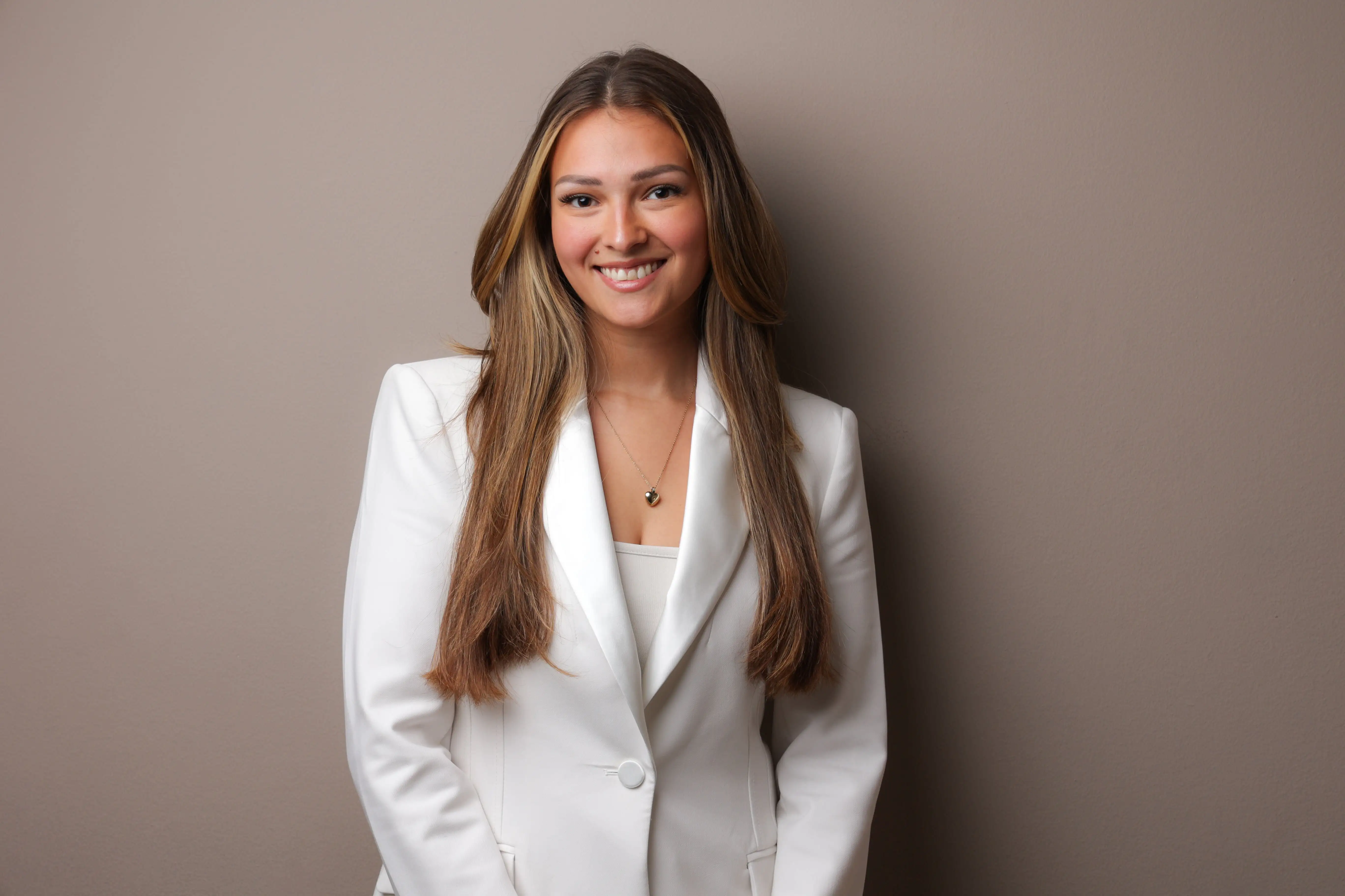 Smiling woman with long hair wearing a white blazer and heart necklace against a beige background.