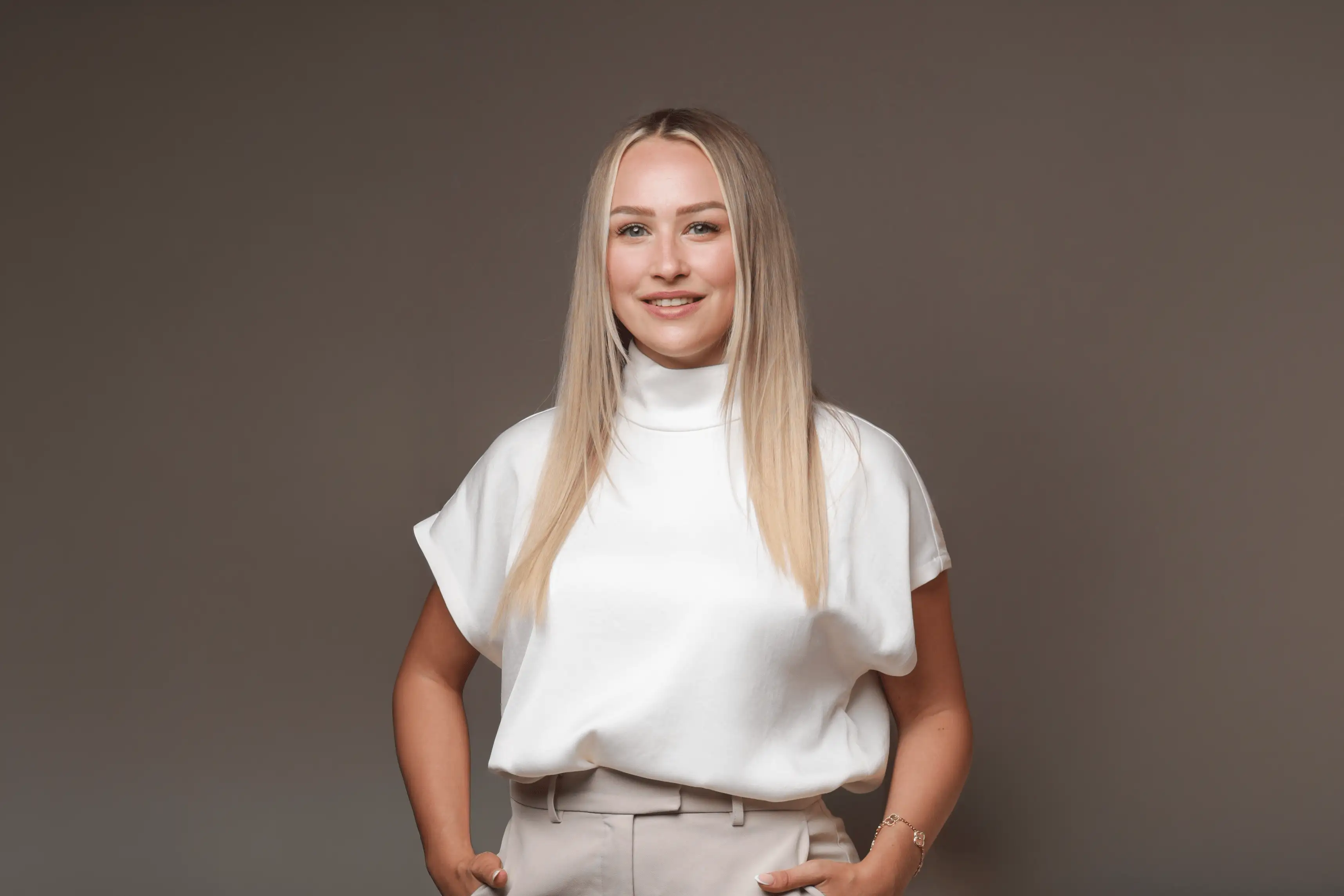 Smiling woman with long blonde hair wearing a white turtleneck top and beige pants on gray background.