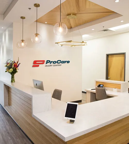 Modern reception area with white and wood-paneled desks, hanging globe lights, a flower vase, and a ProCare Injury Centers sign on the wall.