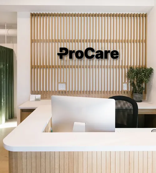 Reception desk with a computer and black chair in front of a wooden slat wall featuring the black ProCare logo and a potted plant on the desk.