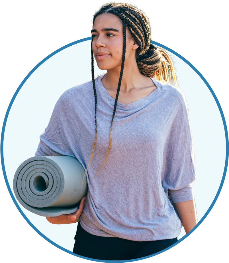 Young woman with braided hair holding a rolled yoga mat, wearing a light gray loose top and black pants.