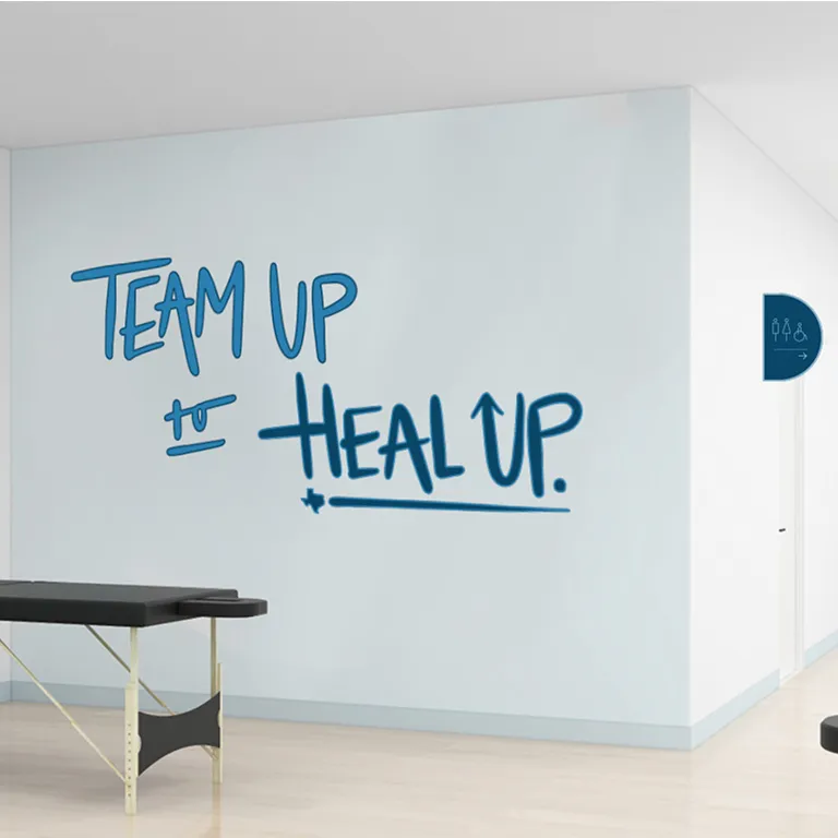 White wall with blue text reading Team up to Heal Up in a medical or therapy room with an examination table.