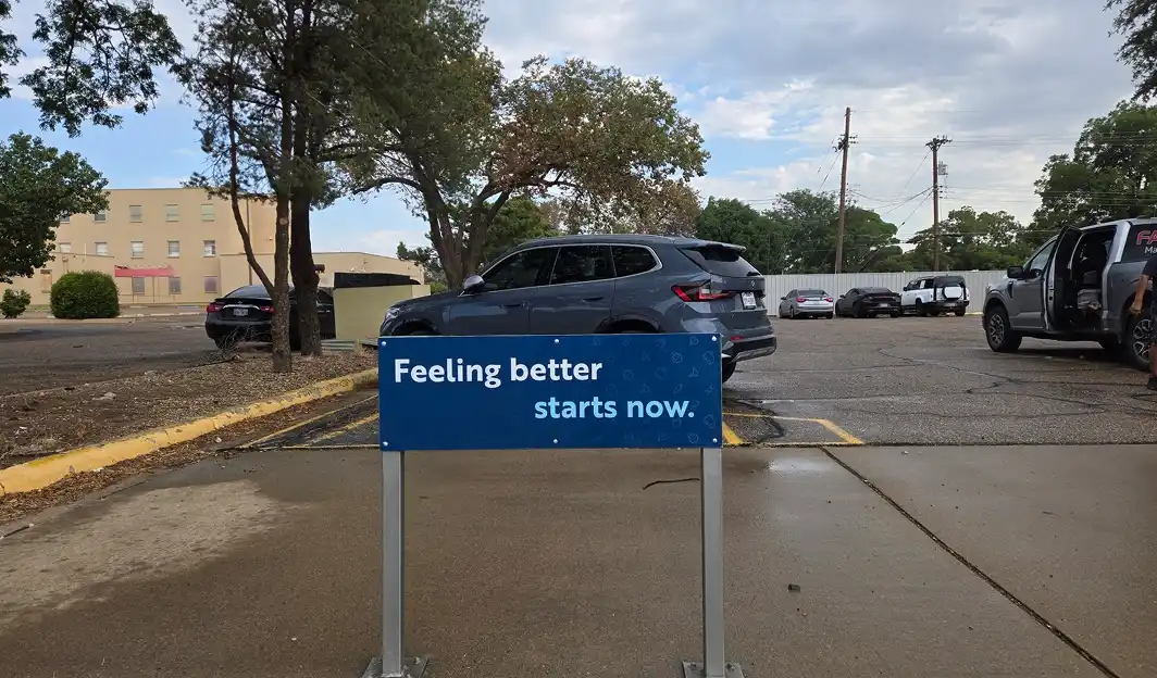 Blue sign with white text that reads 'Feeling better starts now.' in a parking lot with several cars and trees in the background.