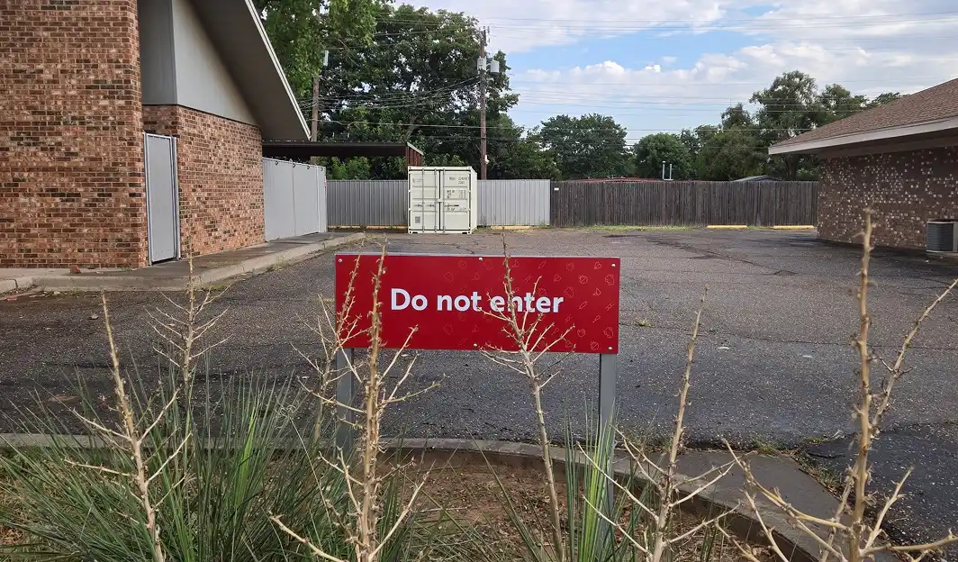 Red sign with white text reading 'Do not enter' in front of a parking area with buildings and a storage container.
