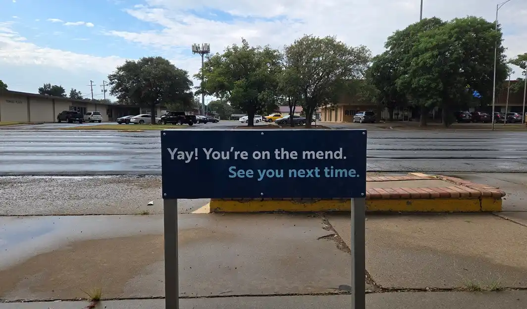 Blue sign on sidewalk reads 'Yay! You're on the mend. See you next time.' with a street and trees in the background.