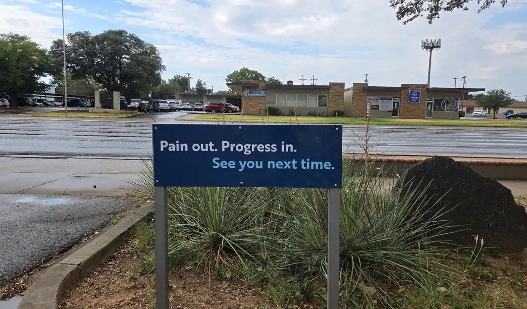 Blue sign with white and light blue text reading 'Pain out. Progress in. See you next time.' placed among plants near a street with commercial buildings in the background.