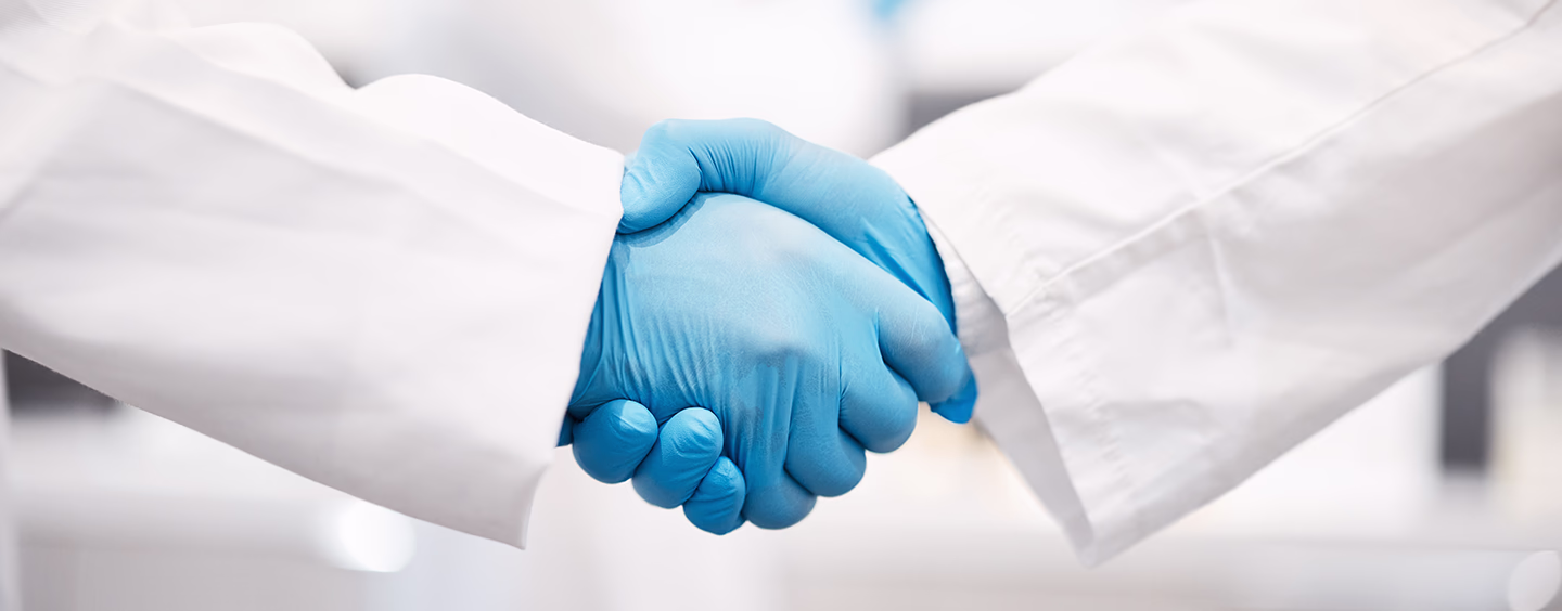 Two lab technicians in white coats and blue gloves shaking hands, close-up crop