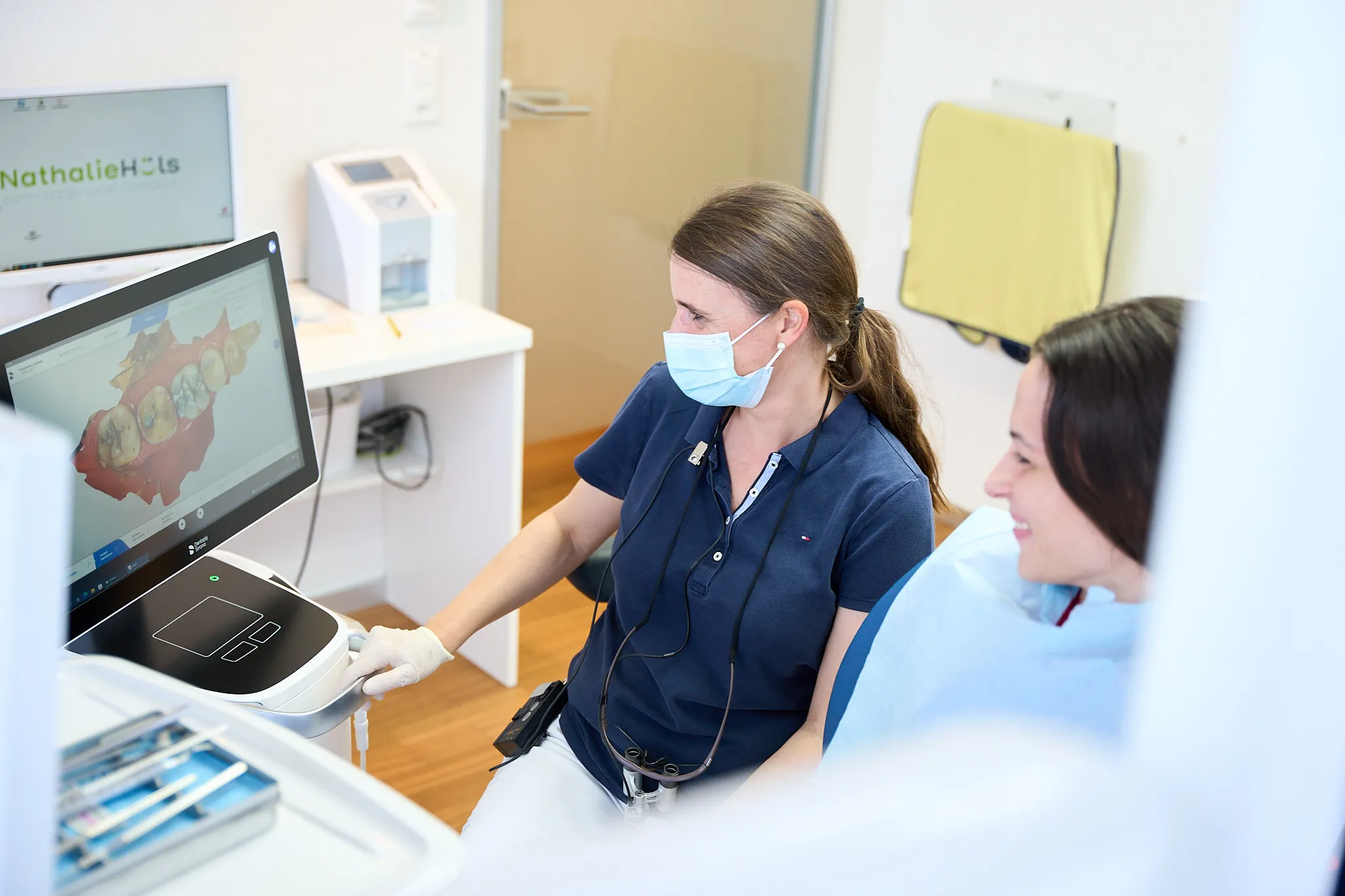 Dentist wearing a mask and gloves showing a 3D dental scan on a monitor to a smiling patient in a dental office.